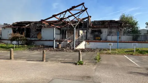 A one-storey white building that has been damaged in a fire. The building has no roof and its internal structure has been exposed.