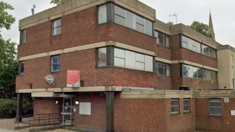 Google Kettering's former police station on London Road. It is a brutalist-style, three-storey red brick building, with large windows on the top floor. It has a "For Sale" board above the front door.