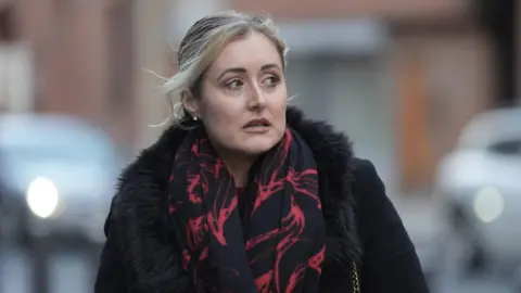 A woman wearing a black coat and a black and red patterned scarf walks through a London street.