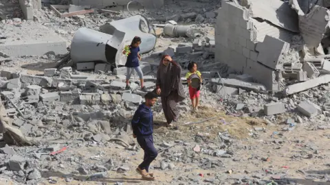 Getty Images Adults and children in Rafah, walking through an area of concrete rubble after an attack by Israel