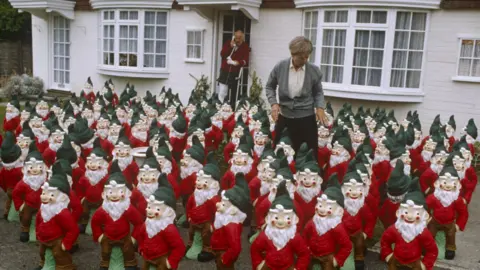 A scene from One Foot in the Grave with the front garden on Victor and Margaret's house covered in garden gnomes. Victor is standing at the front door on the phone and Margaret is in the middle of the gnomes.