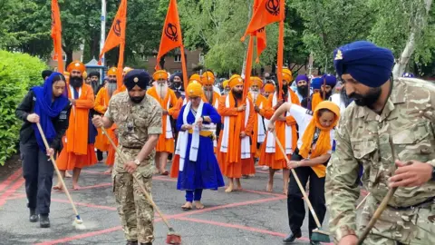 West Midlands Police A group of people take part in a procession on a concrete path flanked by trees. The two front rows are made up of men wearing orange clothes and orange turbans. The five men at the very front are each holding an orange flag.