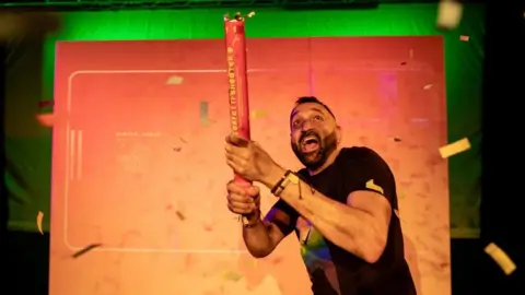A man wearing a black T shirt holding a confetti canon and smiling while he looks into the sky