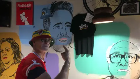 Dieter Steinbusch Dieter pointing at the wall of the pub which has drawings connected to Wrexham. He is pointing at Ryan Reynolds, on the right is Danny DeVito in a nod to a hit show Rob McElhenney stars in. There is also a woman on the left of Dieter drawn on the wall.