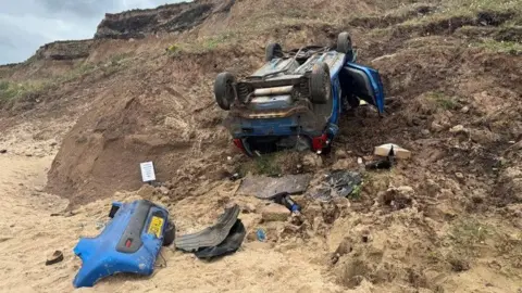 BBC A blue Peugeot 107 car lies upside down at the bottom of a cliff. The back of the car has separated from the rest of the car and parts of the wreckage lies in the sand.