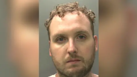 Craig Winston is wearing a white t-shirt. He has short cropped facial hair and curled brown hair.