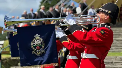 Trumpeters from The Band of the Princess of Wales's Royal Regiment, who are wearing red uniforms and black spiked helmets, playing on their trumpets, which have banners featuring the band's crest hanging from them.