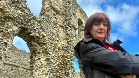 Cosey Fanni Tutti looking down at the camera. She has her arms crossed and is smiling. She is wearing a black coat. Behind her are the ruins of an old priory.