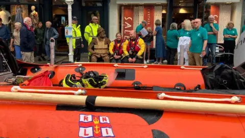 BBC/Sumaiya Motara Two RNLI crew members in full kit are sitting on the bench next to the willow model of an RNLI crewman. There are about five members of the public nearby, along with about seven people in turquoise polo shirts. A lifeboat can be seen in front of the bench. 