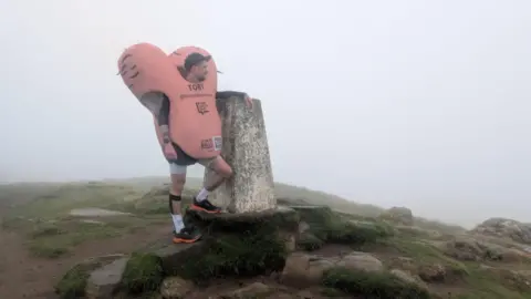 A man dressed in a large pink costume shaped like testicles stands on top of a foggy hill next to a stone marker.