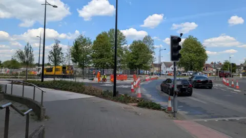 Swindon Borough Council A foot path next to a road where road works are happening. There are a few workers in high vis at the site and two cars are driving past.