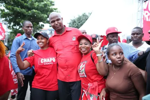 AFP Daniel Chapo merayakan kemenangannya bersama pendukungnya di Maputo pada bulan Desember. Mereka mengenakan pakaian berwarna merah, warna pesta Frelimo-nya. Dua wanita merangkul politisi tersebut dan membuat tanda 'perdamaian' dengan tangan mereka.