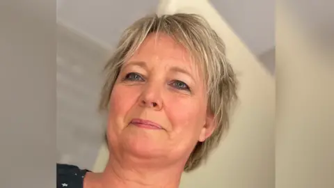 Alison has short blonde hair and blue eyes. She is looking to the left of the camera and smiling. She is stood against a plain cream coloured background.