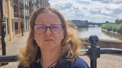 A head and shoulder shot of Su Roys, who has has shoulder-length brown hair, wearing a navy England Rugby polo shirt and burgundy-rimmed glasses. She is stood on a path in front of the River Trent. The bridge can be seen in the distance.