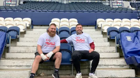 BBC Two men sit in an empty football stadium, surrounded by blue and white seats. They are wearing white tshirts which reads "heartbeat"