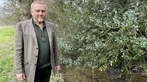 Alex Gater standing next to a leat which has been contaminated by raw sewage. He is looking at the camera and is wearing a grey suit jacket with a green shirt. The leat has stones at the bottom and there is a large branch, with green leaves, hanging over the top of it.