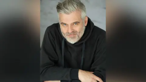 A headshot of a man with silver hair and a beard, smiling at the camera. He wears a black hoodie and has his arms crossed over his chest.