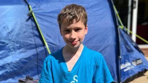 Joshua smiling and wearing  blue t-shirt outside his tent