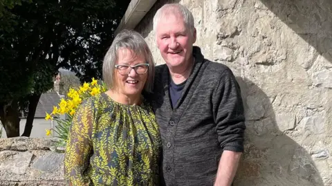 Linda and Alan Boyer smiling in front of a stone wall in the sunshine, both are smiling and Alan has his arm round his wife