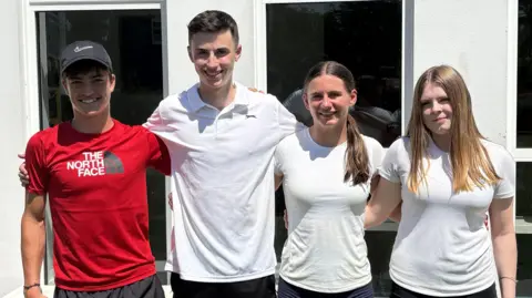 Reece wears a red t-shirt and black baseball cap, Tom has dark hair and a white polo shirt, Cara has brown hair and a white t-shirt, and Maisy blond hair and a white t-shirt. They all stand with arms around one another's backs in front of a school building.