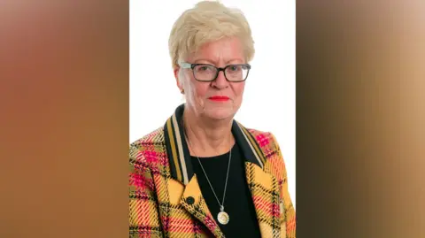 PA Media Chair of the CCRC Helen Pitcher wears a yellow and red jacket over a black top and smiles at camera. She has short blonde hair and glasses 