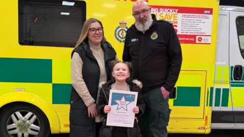 East of England Ambulance Service A little girl grinning while holding a framed certificate in front of her mum and the call handler, behind all of them is a bright yellow and green ambulance