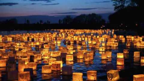 Hundreds of water lanterns float on a lake with trees in the background