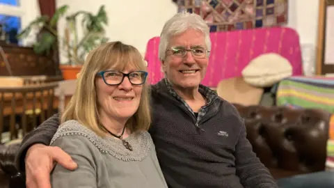 Nikki Little sits on the left wearing a grey top while partner John Porter sits to her right wearing a grey fleece. Both wear glasses an are smiling toward the camera. 