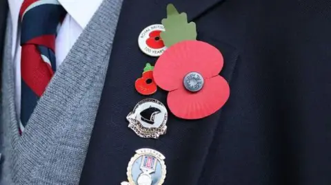 BBC A close-up picture of a man wearing a dark blazer with a red poppy and other military medals on it, The man is also wearing a grey cardigan and a red and blue striped tie
