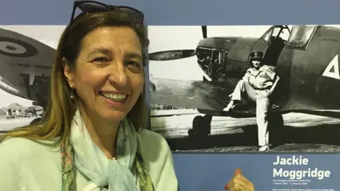 Candy Adkins A smiling Candy Adkins standing in front of a museum display board about her mother with a black and white photo of her posing with a Spitfire