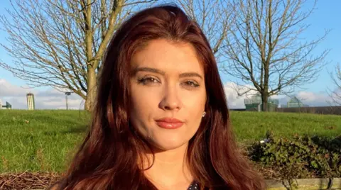 Shows a woman with long dark hair facing the camera and standing in front of a field with three large trees and buildings in the background