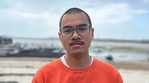 Theo has short hair and glasses. He is wearing an orange sweatshirt standing in front of a sandy shoreline with boats and a pier in the background under a cloudy sky