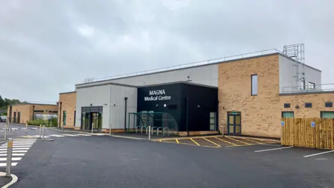 A grey and brick rectangular, flat-roofed building housing a new medical centre, with a car park in front of it. It is a grey, cloudy day.