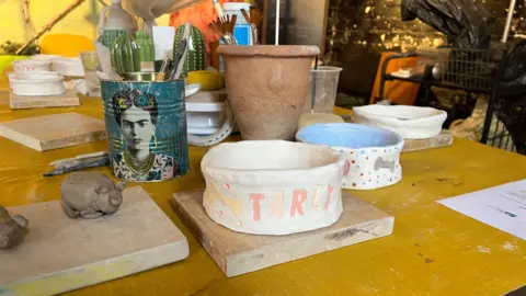 White hand made dog bowl on brown table with pots and other pottery items surrounding it.