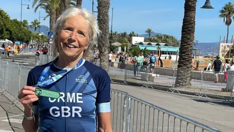 Judy Orme standing at the finish line of the triathlon in Spain. She is wearing a blue cycling jersey which reads Orme and GBR. She is holding up a medal which is also draped around her neck. She is smiling into the camera. Palm style trees are seen in the background.