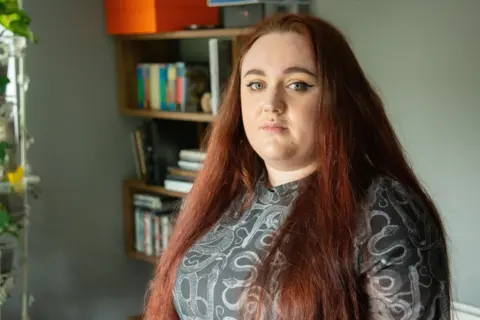 A woman with long dyed red hair and blue eyes stand in her living room.
