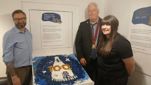 Stoke-on-Trent City Council A man wearing a blue shirt is standing next to a large-scale model of Stoke-on-Trent's centenary logo. It is made up of hundreds of black, blue and white plastic Lego bricks and features the number 100 and the words "Stoke-on-Trent". A man wearing a suit and a woman wearing a black T-shirt are standing on the other side of the display.