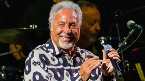 Getty Images Tom Jones standing on stage smiling holding a microphone. There is another musician in the background and drums are visible. 