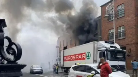 Social Media A still from a video shows a man on top of a Spar lorry. Beside the lorry, is a building on fire with smoke pouring out of the windows. cars and people are visible in the foreground.