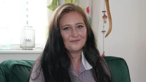 A woman with long dark hair sits on a green sofa. She is smiling at the camera. She wears a shirt which has small blue and red chequers and has a white collar. 