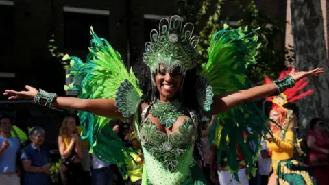 A dancer in green with a flurry of feathers behind her holds her arms out with a wide smile.