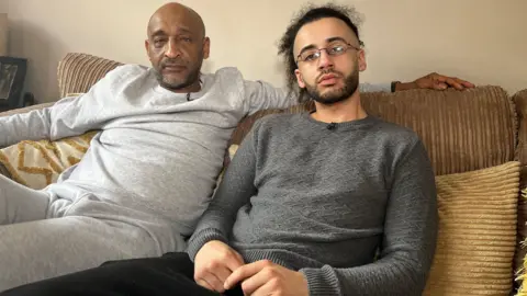 Dean De Freitas and his son Max sit on a beige corduroy sofa together in their home. They are both looking at the camera. Dean is on the left, he is bald with short grey stubble and wears a light grey sweatshirt and jogging bottoms, his arm is resting on the sofa cushion behind Max. Max has black hair which is tied back in a bun and a short black beard. He wears thin wired framed glasses with a dark grey cable knit jumper and black jogging bottoms.