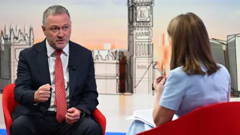 BBC Housing, Communities and Local Government Secretary Steve Reed appearing on the Sunday with Laura Kuenssberg, with presenter Victoria Derbyshire standing in. The pair sit on red bucket chairs. Reed wears a black suit with a white shirt and a red tie. Derbyshire is wearing a pastel blue dress with short sleeves. She has shoulder length, dark blonde hair. 