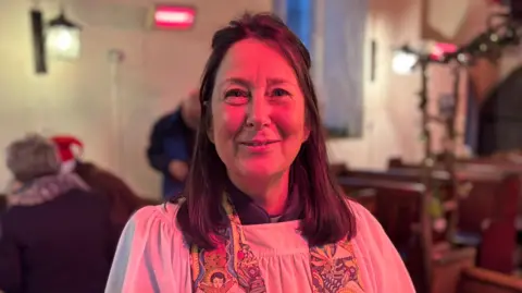 Luke Deal/BBC Reverend Emma Barr standing between pews inside St John the Baptist Church in Mount Bures, near Colchester. She has brunette hair and is looking directly at the camera and smiling. 