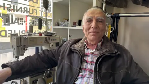 Jo Burn/BBC A man with short, grey hair sat beside a window and a large white sewing machine. He is wearing a checked shirt and a brown jacket.