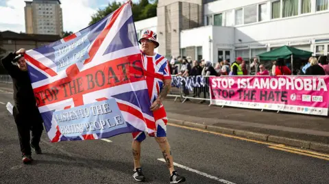 Getty Images Anti-migrant protesters outside the Cladhan Hotel on 13 September. Two men carry a union flag with the phrase Stop the boats written on it. The man at the back is dressed in black and the man at the front is wearing shorts, a tshirt and a hat which all feature the flag. In the background are counter-protesters and a banner reading: Stop Islamophobia. Stop the hate.