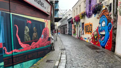 A wide picture of Mansell Street. It has a cobbled road, and the walls are painted with vibrant colours. At the end of the street is a road sign to direct people into the gallery building.