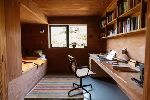IWC Media A compact, wood-panelled room features a built-in bed on the left with cushions and a mustard-coloured blanket.
Opposite, a long wooden desk holds an open laptop, books, and small objects, with shelves above filled with neatly arranged books and framed photos.
A window centred on the back wall lets in daylight and reveals a grassy outdoor scene, while a striped rug and simple chair complete the cosy workspace.