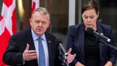 Danish Foreign Minister Lars Lokke Rasmussen wears a black suit and is gesturing with his right hand as Greenland's Foreign Minister Vivian Motzfeldt stands next to him wearing a navy jacket and looking downwards