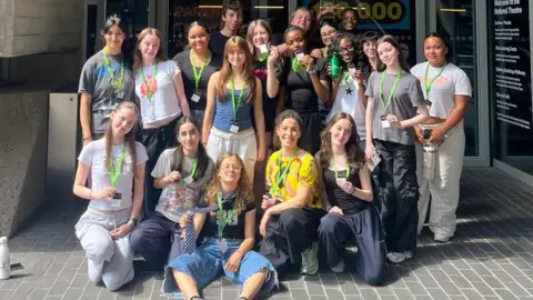 A group of teenagers standing outside the National Theatre in London. The teenagers are standing in rows, with those at the front kneeling or sitting down. They are wearing casual clothes.